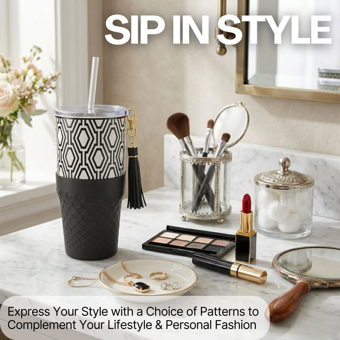 Tabletop scene with a Biddlebee 30oz Black & White Charm tumbler with a straw lid, vegan tassel and silicone boot on a white background, makeup items, and jewelry with 'Sip in Style' text.