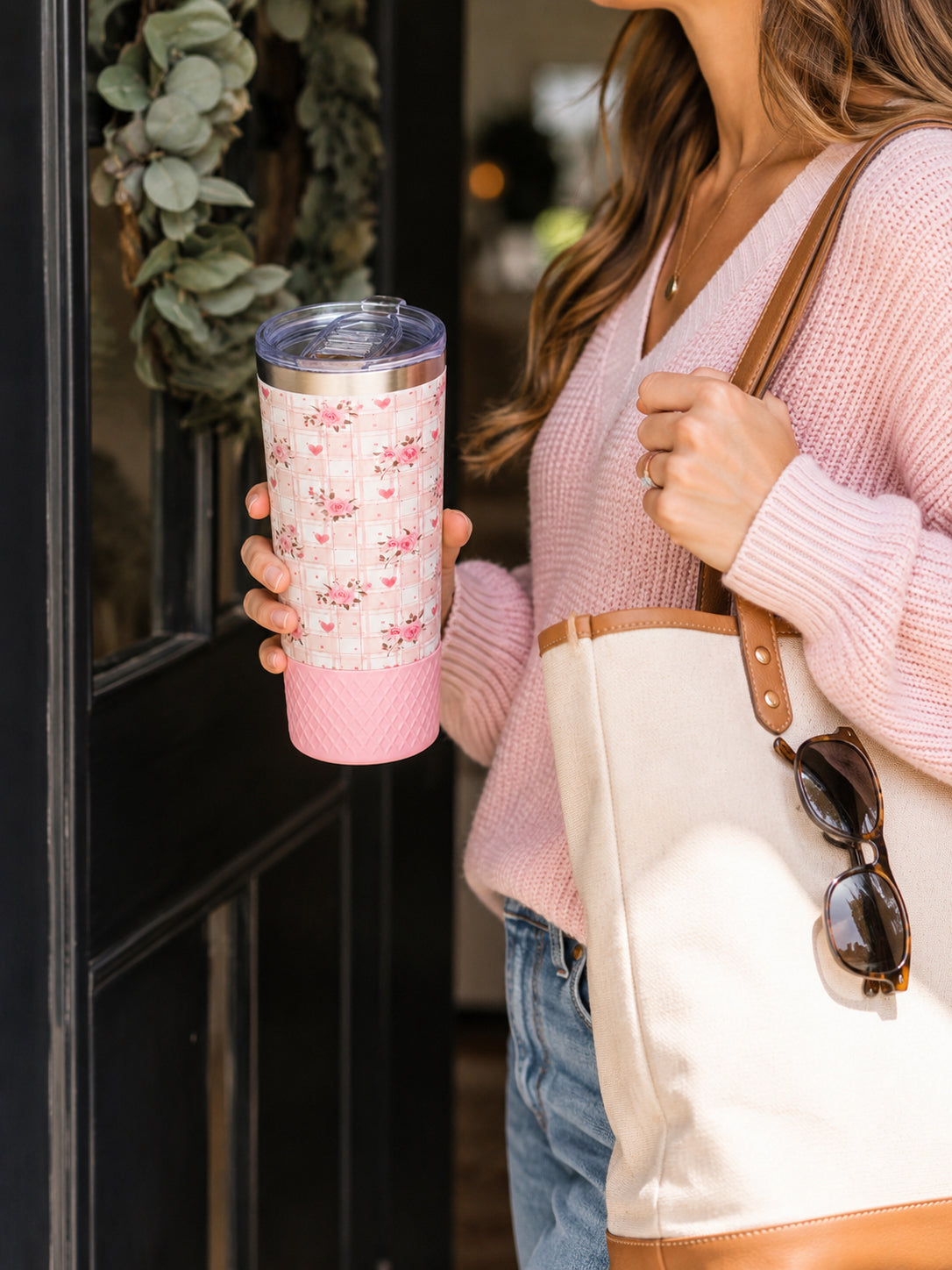Woman in a pink sweater holding a Biddlebee 20oz Signature Collection tumbler with a pink floral heart plaid print and pink silicone boot, fitted with the Biddlebee Brew coffee lid, standing at a front door with a tote bag